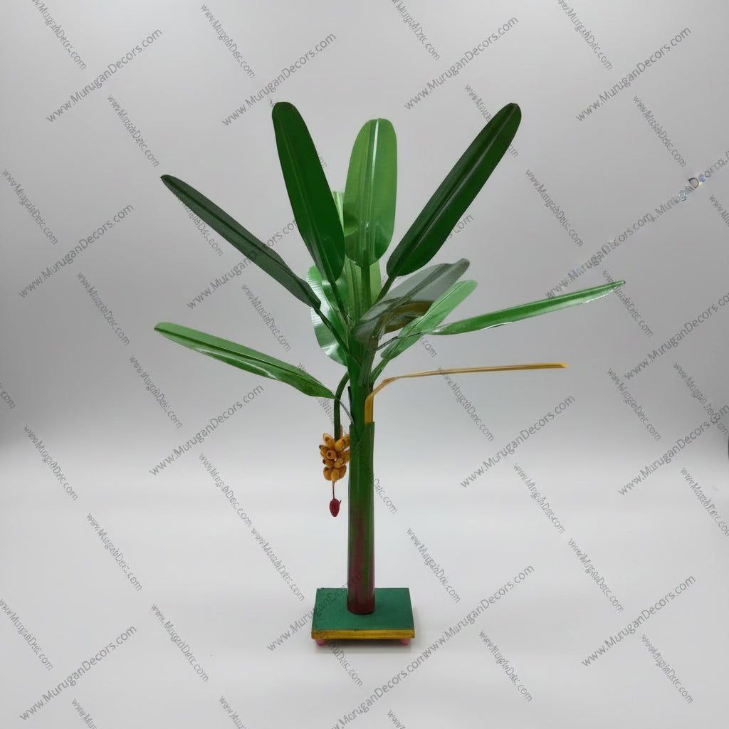 A small potted artificial banana tree plant with green leaves and a brown trunk, displayed on a white table in front of a wooden door.