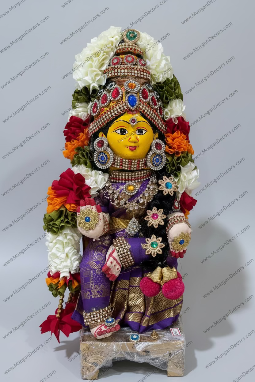 A decorated Hindu deity idol, likely Lakshmi, adorned with colorful artificial flowers, intricate rhinestone jewelry, and a gold-toned crown, standing next to brass oil lamps on a beige wall.
